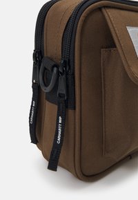 Brązowa torba z materiału z czarnymi zamkami błyskawicznymi i uchwytami, posiadająca teksturowaną powierzchnię i boczną kieszeń, oznakowana marką "CARHARTT WIP."