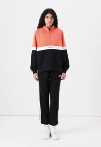 Pullover color block Roxy in corallo e nero con zip, abbinato a pantaloni neri con strisce laterali bianche e logo, indossato con sneakers bianche.