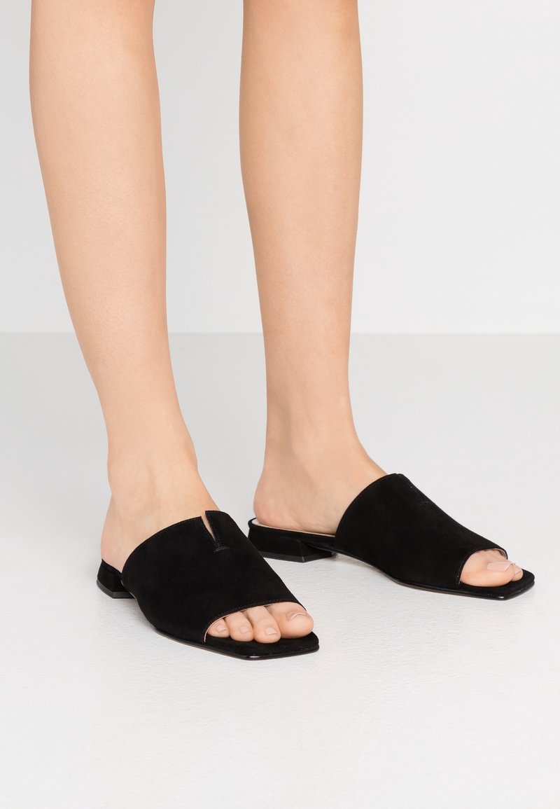 Jambes portant des sandales mules noires à bout ouvert, à enfiler, avec des petits talons blocs sur un fond clair uni.