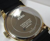 Montre en acier inoxydable doré avec des détails sur le modèle et la résistance à l'eau au dos. Présente un bracelet texturé noir et un logo gravé.
