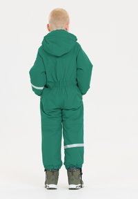 Grøn vandtæt jumpsuit med hætte, elastisk talje og reflekterende striber. Bærers med grå støvler, der har teksturerede såler og lukkedetaljer.