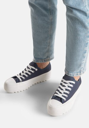 Osoba ubrana w jasnoniebieskie dżinsy i granatowe trampki na platformie z białymi sznurowadłami i grubymi białymi podeszwami stojąca na szarej powierzchni.