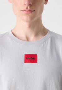 T-shirt in cotone grigio con un'etichetta rettangolare rossa contenente la parola "HUGO" in testo nero. Design a collo rotondo con texture liscia.