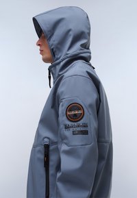 Grau gefütterte Jacke mit glatter Textur, ausgestattet mit einer Reißverschlusstasche, einem runden Logo-Patch und auffälliger Markenkennzeichnung am Ärmel.