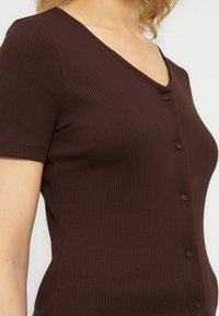 Γυναίκα που φοράει ένα σκούρο καφέ κοντομάνικο πλεκτό τοπ με κουμπιά και V-λαιμόκοψη σε απλό ανοιχτό φόντο.