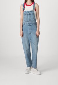 Denim overalls in light blue, featuring adjustable straps, front pockets, and a relaxed fit. Paired with a white and red tank top and white sneakers.