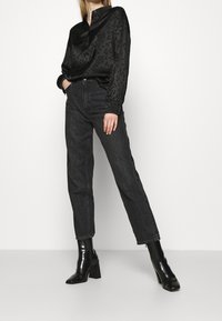 Svart satin skjorta med prickig mönster, i kombination med hög midja mörka denimjeans. Modellen bär svarta klackboots. Enkla vita bakgrund.