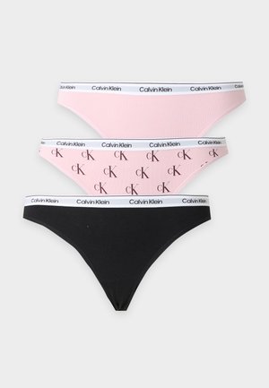 Femme debout pieds nus portant un bralette blanc et un sous-vêtement noir avec le logo Calvin Klein sur les bandes élastiques, sur un fond clair uni.