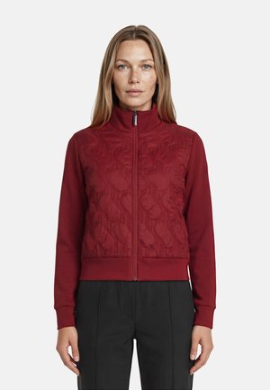 Vrouw met steil blond haar, gekleed in een rood gewatteerd jasje met rits en zwarte broek, staand tegen een effen lichte achtergrond.