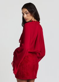 Blusa rossa semi-trasparente con maniche a volants, caratterizzata da un colletto arricciato. Abbinata a una mini gonna rossa strutturata decorata con motivi simili a rose.