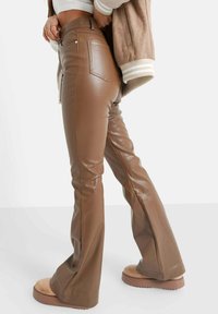 Bruine neplederen flared broeken met een hoge taille, voorzien van twee achterzakken en een gladde, glanzende textuur. Gemaakt in combinatie met lichtgekleurde schoenen.