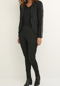 Veste noire cintrée avec un seul bouton, associée à un pantalon noir fuselé avec une discrète bande latérale. Les deux réalisés en tissu lisse.