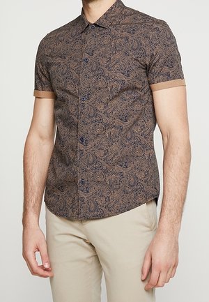 Mężczyzna noszący krótką koszulę na guziki z brązowym wzorem paisley i beżowymi mankietami, w połączeniu z jasnobeżowymi spodniami.
