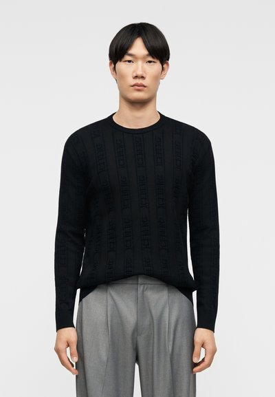 Jeune homme debout, portant un pull noir à manches longues avec des motifs subtils de texte vertical et un pantalon plissé gris clair.