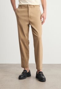 Chino kaki avec une coupe droite, en coton léger, dotés de poches latérales et d'une fermeture à bouton. Associés à des mocassins noirs.