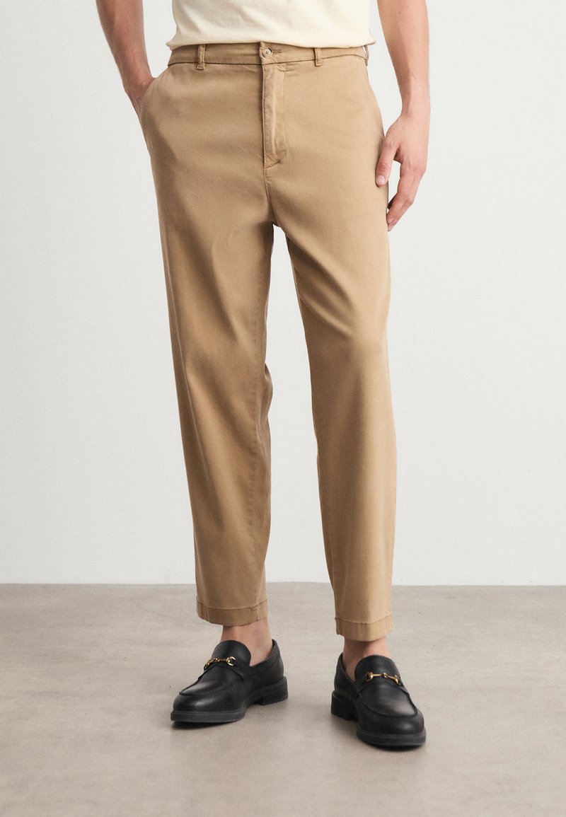 Chino kaki avec une coupe droite, en coton léger, dotés de poches latérales et d'une fermeture à bouton. Associés à des mocassins noirs.