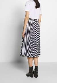 Kvinna iklädd en svartvitt randig plisserad midikjol, vit kortärmad topp och svarta nitar ankle boots, stående mot en vit vägg.