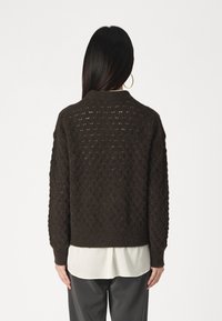 Pull en tricot marron foncé présentant un motif texturé en losanges, un col rond et des poignets côtelés, porté sur un chemisier blanc.