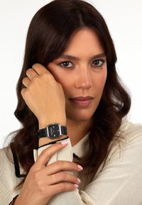Orologio rettangolare nero con accenti in argento, cinturino nero texturizzato. Modella che indossa un top beige chiaro con bordi neri, mentre mostra l'orologio.