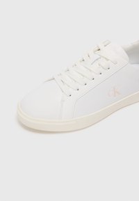 Fehér, alacsony szárú sneaker, sima szintetikus anyagból készült, lapos fehér fűzőkkel, diszkrét dombornyomott logóval és kerek orral.