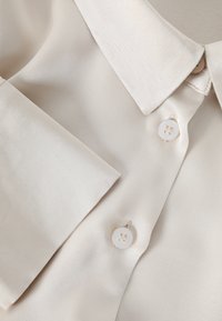 Bézs szatén blúz részlete, hegyes gallérral, zsebbel és két finoman fénylő gombbal. Sima textúra, szabott kialakítás.