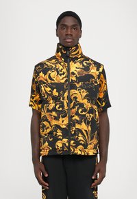 Un jeune homme portant un gilet matelassé noir et doré à motifs floraux avec un col haut et une chemise à manches courtes assortie, se tenant devant un fond blanc.