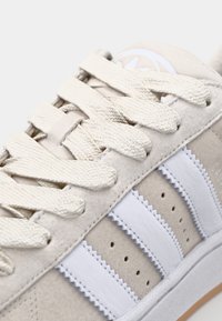Beige suedesneaker med hvite lærstriper, teksturerte snorer og kremfargede lisser. Har en buet såle og minimal merkedetalj.