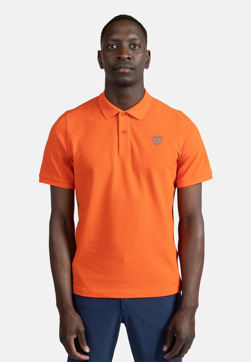 Oranžové polo tričko vyrobené z bavlny s klasickým límečkem, dvěma knoflíky a malým logem na levé hrudi. Má krátké rukávy.