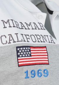 Vitt polo shirt med grått tygpanel som har "MIRAMAR CALIFORNIA" i grå broderi och en broderad amerikansk flagga med "1969" i blått.