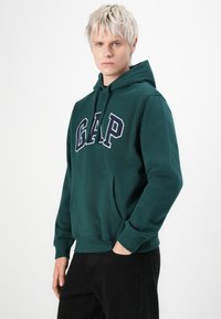 Young person with short blond hair wearing a dark green GAP hoodie and black pants, standing with one hand in pocket.