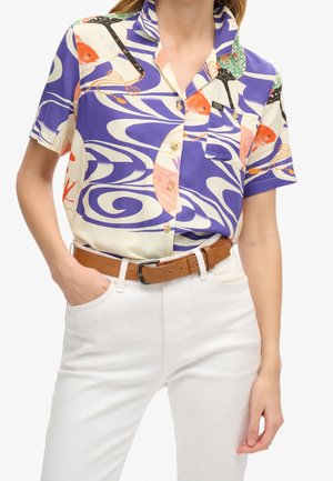 Femme portant une chemise à manches courtes avec un motif abstrait de poissons violet, blanc et orange, pantalon blanc et ceinture marron.