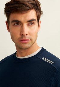 Sweatshirt bleu marine texturé avec un tissu côtelé, encolure arrondie et un détail blanc au niveau du col. Logo "FREDDY" sur l'épaule.