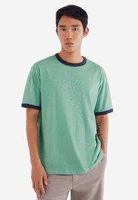 Grøn kortærmet t-shirt med marineblå kant, rund halsudskæring, afslappet pasform og subtil teksturmønster; iført med lysegrå bukser.