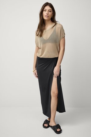 Top en tricot beige à coupe décontractée, jupe maxi drapée noire avec fente latérale, et sandales noires avec des brides nouées.