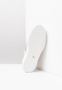 Baskets avec une semelle blanche en caoutchouc et des accents métallisés beiges. La tige présente des textures lisses et un logo minimaliste.