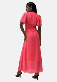 Robe maxi en rose vif, avec des manches courtes, une taille smockée et un tissu transparent avec une texture lisse. Sandales à talons hauts noires.
