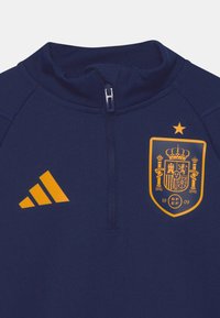 Marinblå sportjacka med hög krage, kvartdragkedja, orange Adidas-logotyp och Spaniens landslagets emblem med en krona och stjärna.