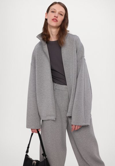 Pelēks zip-up sporta džemperis ar augstu apkakli, kombinēts ar atbilstošām sporta biksēm. Gluda tekstūra, oversize griezums un sānu kabatu detaļas. Melna rokassoma.