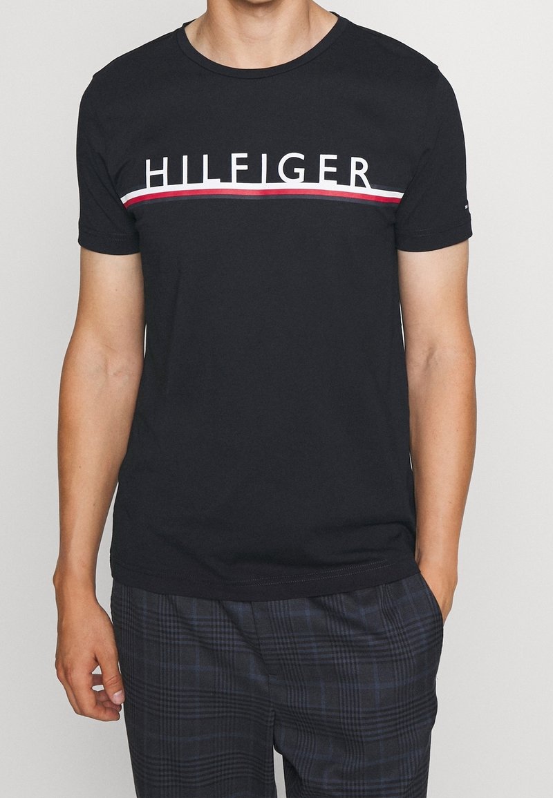 Muž v černém tričku s nápisem "HILFIGER" a červenými a bílými pruhy přes hruď, doplněný tmavými kostkovanými kalhotami.