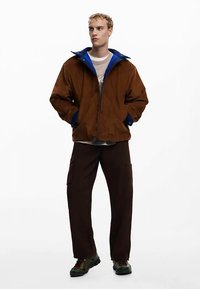 Bruine jas met ritssluiting en een blauwe kraag, gedragen over een grijs shirt. Gecombineerd met donkerbruine cargobroek en meerkleurige schoenen.