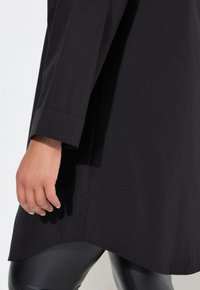 Sort lang tunika skjorte med lige snit, der har en afrundet kant og opbrettede ærmer. Kombineret med skinnende sorte leggings.