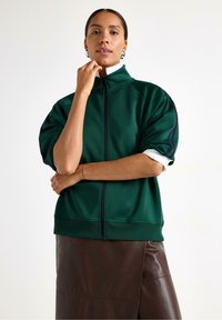 Grøn zip-up sweatshirt med høj krave, oversized korte ærmer og navy detaljer, parret med en mørkebrun lædernederdel.