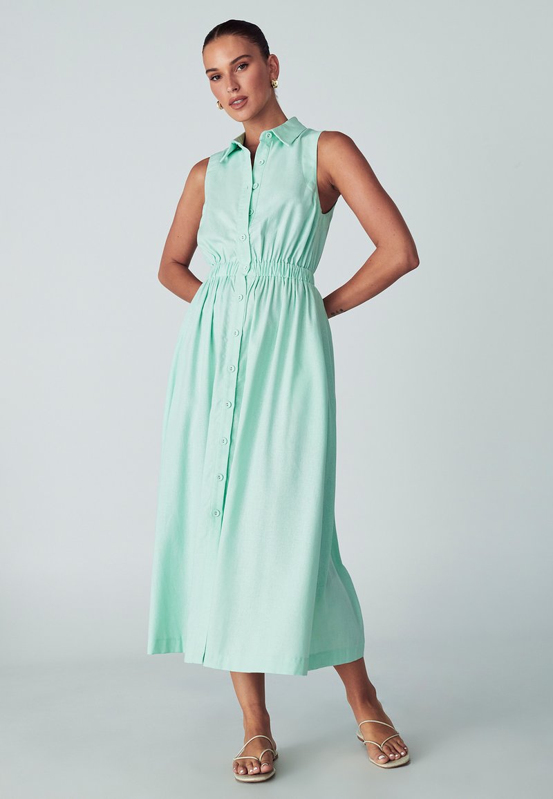 Šviesiai mėtų žalios spalvos be rankovių suknelė su apykaklės iškirpte, mygtukų uždarymu priekyje ir elastiniu juosmeniu, suteikiančiu aptemptą siluetą. Lygus audinio paviršius.