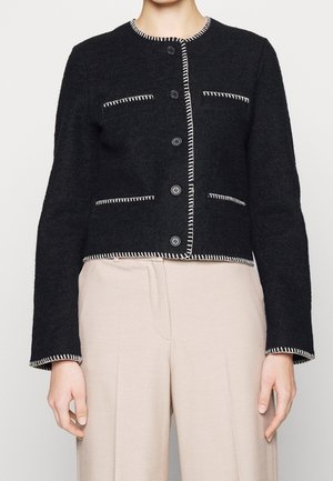 Veste cropped en mélange de laine noire avec un col rond, mettant en avant des coutures blanches le long des bords et six boutons à l'avant, assortie à un pantalon beige.