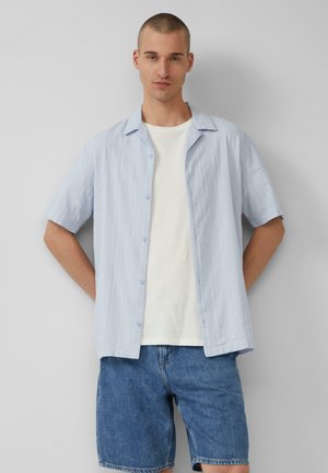 Lichtblauw overhemd met korte mouwen, met een knoopsluiting aan de voorkant, een gestructureerde stof en een klassieke kraag, gedragen over een wit T-shirt en denimshorts.