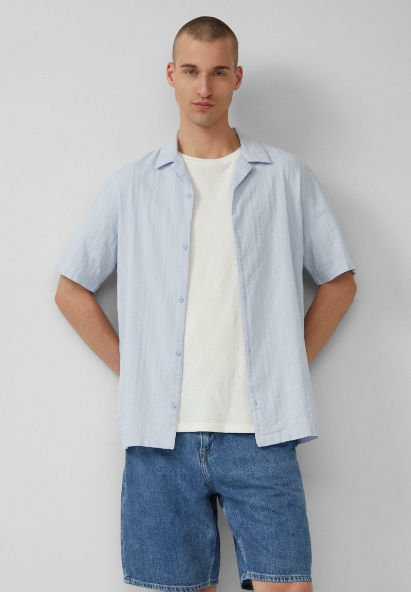 Lichtblauw overhemd met korte mouwen, met een knoopsluiting aan de voorkant, een gestructureerde stof en een klassieke kraag, gedragen over een wit T-shirt en denimshorts.