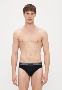 Juodos medvilninės trumpikės su pilku elastiniu liemeniu, ant kurio juodais rašmenimis pavaizduotas prekės ženklo pavadinimas „BOSS“. Lygus paviršius, glaustinantis dizainas.