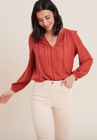 Blouse couleur rouille avec un motif rayé texturé, encolure en V et manches longues. Associée à un jean ajusté beige clair.