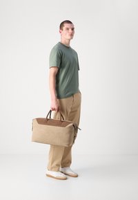 Sac de voyage beige avec des accents en cuir marron, fermeture éclair et doubles poignées. Le modèle porte une chemise verte et un pantalon beige, se tenant sur un fond blanc.