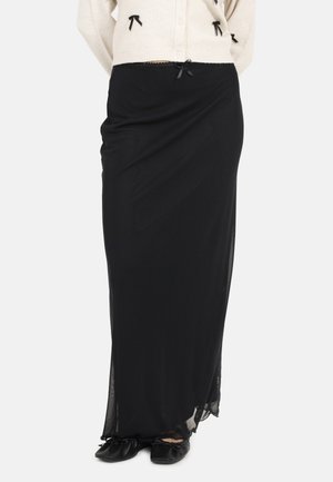 Vrouw die een lange zwarte rok draagt met een klein strikje in de taille en zwarte platte schoenen, gecombineerd met een crèmekleurige trui met kleine zwarte strikjes.